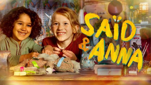 Saïd sitzt links neben Anna an einem Tisch. Beide lachen und halten Bastelutensilien in den Händen. Rechts steht der Titel Saïd und Anna.