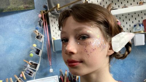 Ein Porträt von Julia. Sie ist mit Glitter auf Wangen und Nase als Elfe geschminkt. Ihre Haare sind geflochten und hochgesteckt. Im Hintergrund eine blaue Wand mit einer Fotogirlande und einem weißen Schreibtischboard.