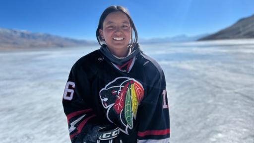 Ein Mädchen mit dunklen Haaren steht in Eishockeyausrüstung auf einem zugefrorenen See und lächelt.