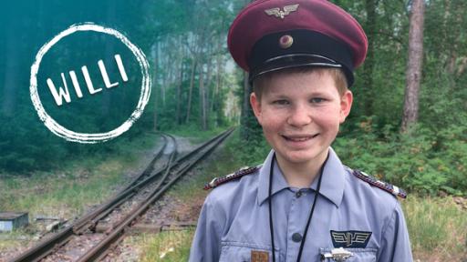 Willi steht in Lokführer-Uniform an einer Gleisanlage im Wald; Text im Bild: WILLI (Quelle: rbb)