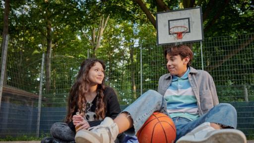 Ein Mädchen und ein Junge sitzen untrer einem Basketballkorb und lächeln sich an.