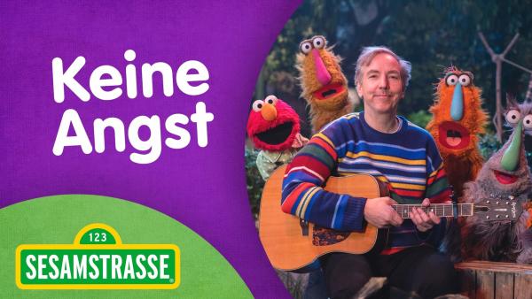 Musiker Olli Schulz sitzt mit einer akustischen Gitarre und Elmo und drei Puppen namens Lankies sitzen hinter ihm und schauen über seine Schultern.