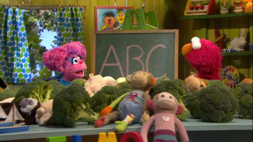 Abby, das pinke Monstermädchen steht links und Elmo, der rote Monsterjunge rechts vor einer Tafel auf der ABC steht.