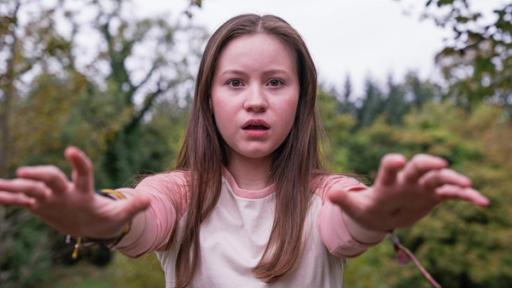 Meg, gespielt von Katy Byrne , steht draußen vor Bäumen und Büschen. Der Wind weht ihr ins Gesicht und sie schaut angestrengt nach vorne. Ihr Blick sieht besorgt aus.