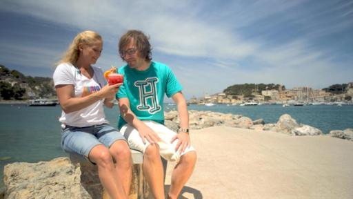 Singa und herrH sitzen auf einem Stein einer Hafenpromenade. Singa, die ein weißes SingAlarm-Shirt und eine kurze Jeans trägt, hält einen roten Cocktail in der Hand. Ihr Urlaubsgast herrH, der ein türkises herrH-Shirt und eine weiße Hose trägt, sieht auf den Cocktail.