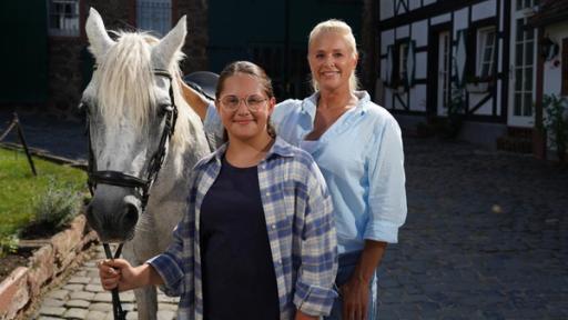  Verhexte Reiterferien wie bei Bibi & Tina": Maya und Singa stehen im Innenhof eines Reiterhofes. Maya hält das Pony Anouk am Zügel, das von Singa gestreichelt wird.