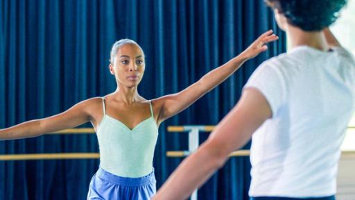 Cece hat ihr Trainingsballett-Outfit an und nimmt eine Tanzpose ein. Ihr linker Arm ist erhoben. Benoit steht ihr gegenüber. Cece schaut angestrengt.