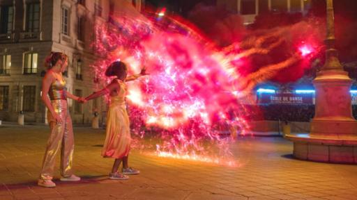 Abends auf einer Straße in Paris: Jade und Cece werden von dunkler Mystic-Magie angegriffen. Cece wehrt diese mit Wizen-Kräften ab. Ein rot glühender Schutzwall baut sich vor ihnen auf. Jade steht hinter Cece und hält deren Hand fest.