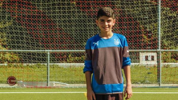 Ayham steht auf dem Fußballplatz vor dem Tor. Er trägt ein blau/schwarzes Trikot und schaut freundlich in die Kamera. Die Sonne scheint. | Rechte: ZDF