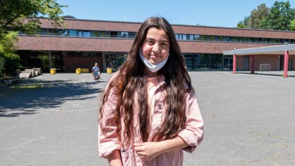 Azra steht mit heruntergezogener Maske vor dem Schulgebäude. Azra hat langes dunkles Haar, trägt eine Jeans und eine rosafarbene Jacke. Der Schulhof hinter ihr ist leer. Sie lacht in die Kamera. | Rechte: ZDF