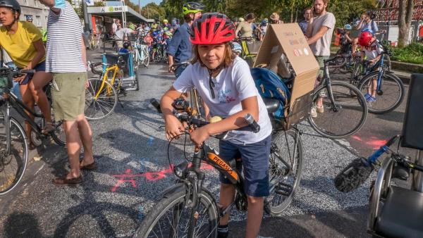 Jupp trägt einen roten Fahrradhelm und steht an seinem Rad mitten unter vielen Radaktivisten bei der Fahrraddemo. Er trägt ein weißes T-Shirt mit Aufschrift.  Die Sonne scheint. Jupp lächelt in die Kamera. | Rechte: ZDF