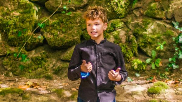 Tiago steht vor einer moosbewachsenen Felswand im Wald. Hier will er seine erste Unterrichtsstunde halten. Er trägt eine Jeans und ein dunkles Hemd und wirkt konzentriert und zuversichtlich. | Rechte: ZDF