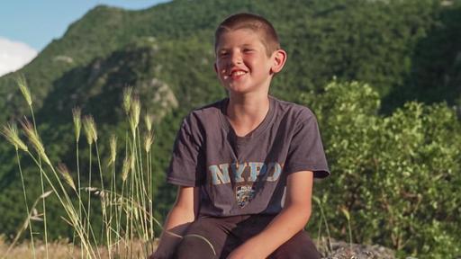 Ilija sitzt auf einem Felsen und lächelt fröhlich in die Kamera. Die Sonne scheint. Im Hintergrund kann man einen bewaldeten Hang erkennen. Alles wirkt sehr ruhig und friedlich.