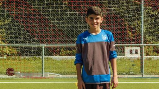 Ayham - Anpfiff in mein neues Leben | Rechte: ZDF Ayham steht auf dem Fußballplatz vor dem Tor. Er trägt ein blau/schwarzes Trikot und schaut freundlich in die Kamera. Die Sonne scheint.