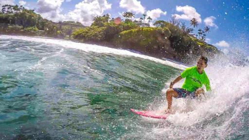 Diesel trägt ein neonfarbenes Surfshirt, dunkle Surfshorts und surft eine Welle. Das Wasser spritzt hinter ihm auf. Im Hintergrund ist das Ufer zu erkennen.