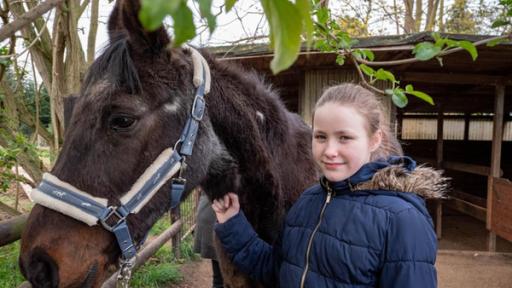 Evi steht mit ihrem alten Pferd Foxi vor dem Stall. Evi hat eine blaue Jacke an mit Fellkragen. Ihr blondes langes Haar hat sie zu einem Zopf gebunden. Sie lacht in die Kamera. Mit der rechten Hand hält sie ihre Pferd Foxi am Halfter.