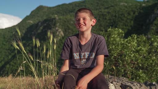 Ilija sitzt auf einem Felsen und lächelt fröhlich in die Kamera. Die Sonne scheint. Im Hintergrund kann man einen bewaldeten Hang erkennen. Alles wirkt sehr ruhig und friedlich.