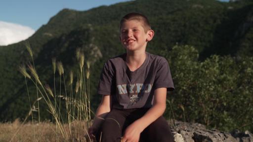 Ilija sitzt auf einem Felsen und lächelt fröhlich in die Kamera. Die Sonne scheint. Im Hintergrund kann man einen bewaldeten Hang erkennen. Alles wirkt sehr ruhig und friedlich.