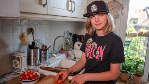 Jakub hat schulterlanges, blondes Haar und eine schwarze Kappe auf. Er steht in der Küche an einem großen Holzschneidebrett und schneidet Erdbeeren. Neben ihm steht eine große Schüssel mit bereits geschnittenen Erdbeeren. Er schaut in die Kamera und lächelt.