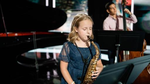  Kika mit ihrem Saxophon in einem Konzertsaal. Sie bläst in das Instrument und schaut dabei konzentriert auf den Notenständer vor ihr. Sie trägt ein festliches blaues Kleid mit Glitzereffekt und einen Haarreif mit Strass-Besatz.