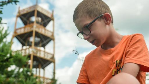 Auf dem Bild ist ein Junge mit orangenem Shirt und Brille der nach unten Schaut. In der Ferne sieht man einen Turm. 