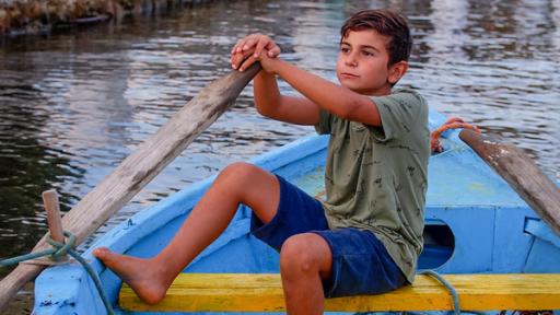 Savvas sitzt in einem kleinen Kahn, der offensichtlich schon lange benutzt wurde, denn seine blaue und gelbe Farbe blättert ab. Savvas hat die Hände auf einem Ruder abgelegt, das mit einem Seil am Boot befestigt ist. Er schaut gedankenverloren in die Ferne. Die Abendstimmung am Meer ist sehr ruhig und friedlich.