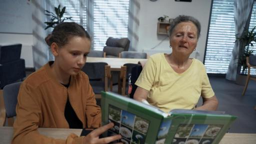 Veronika und ihre Oma sitzen an einem Tisch. Veronika hält ein Buch mit Bildern aus der Natur in der Hand, das sie der Oma zeigt. Im Gesicht der Oma sind deutlich die Spuren der Demenz zu erkennen. Der Raum, den man im Hintergrund wahr nimmt, ist ein mit Sitzmöbeln ausgestatteter Aufenthaltsraum im Altersheim. Alles strahlt Ruhe aus und man erahnt das innige Verhältnis zwischen Oma und Enkelin.