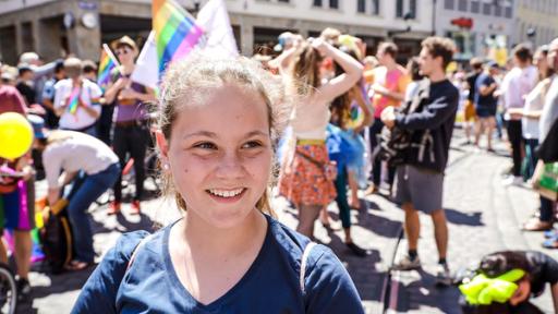 Jane lächelt und blickt an der Kamera vorbei. Im Hintergrund sind viele Menschen in bunter Kleidung und vereinzelt mit Fahnen ausgestattet auf der Straße versammelt.