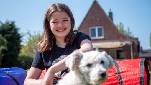 Jule hockt mit dem weißen Hund Bounty auf der Wiese und lächelt in die Kamera. Die Sonne scheint. Es sind Tunnel im Hintergrund zu sehen, durch die der Hund laufen soll.