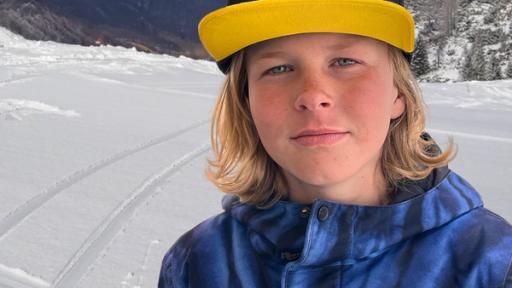 Lan schmunzelt in die Kamera. Er hat blaue Augen, Sommersprossen und langes, blondes Haar. Er trägt eine blaue Winterjacke und eine Baseballmütze mit gelbem Schild. Im Hintergrund sieht man eine verschneite Piste