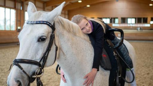 Lenya liegt gemütlich auf dem Rücken ihres Pferdes und lächelt in die Kamera. Dabei umarmt sie ihren Schimmel, Santana, liebevoll. Lenya trägt ihr langes, blondes Haar zum Zopf. Die Reithalle im Hintergrund ist leer.