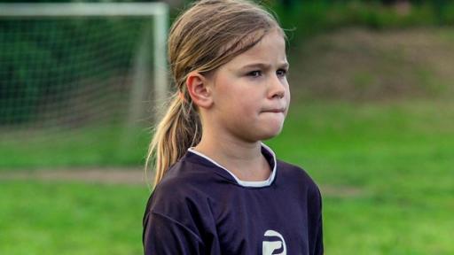 Porträt von Luana. Luana trägt ein Fußball-Trikot und befindet sich auf dem Fußballplatz.  Sie beißt sich auf die Unterlippe und wirkt sehr konzentriert. Im Hintergrund steht ein Fußballtor.