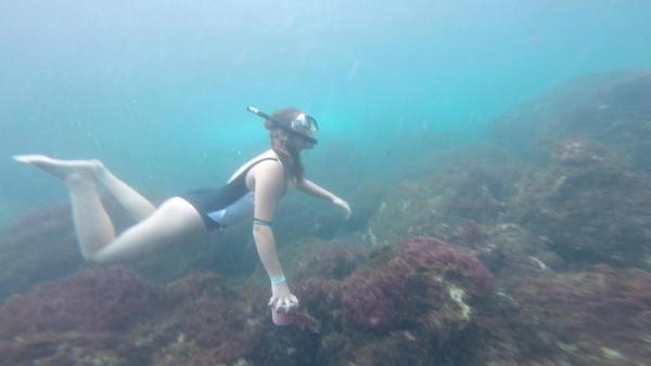 Maria taucht im Meer. Sie trägt einen Badeanzug, eine Taucherbrille und einen Schnorchel. Sie schwimmt über ein Korallenriff. In der Hand hält sie Plastikmüll, den sie gerade vom Boden aufgesammelt hat.