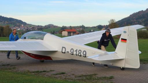 Marius bringt zusammen mit seinem Fluglehrer seinen Flieger zur Startbahn. Sein Trainer schiebt den vordereren Teil des Flugzeugs, während Marius das Heck anschiebt. Im Hintergrund ist eine Berglandschaft zu sehen.