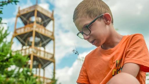 Auf dem Bild ist ein Junge mit orangenem Shirt und Brille der nach unten Schaut. In der Ferne sieht man einen Turm. 