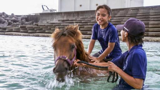  Uruné reitet auf dem Pony im Wasser, der Trainer hält die Zügel des Ponys in der Hand. Uruné lacht glücklich den Trainer an.