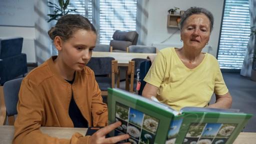 Veronika und ihre Oma sitzen an einem Tisch. Veronika hält ein Buch mit Bildern aus der Natur in der Hand, das sie der Oma zeigt. Im Gesicht der Oma sind deutlich die Spuren der Demenz zu erkennen. Der Raum, den man im Hintergrund wahr nimmt, ist ein mit Sitzmöbeln ausgestatteter Aufenthaltsraum im Altersheim. Alles strahlt Ruhe aus und man erahnt das innige Verhältnis zwischen Oma und Enkelin.