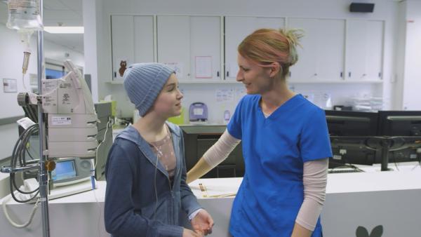 Jenny steht neben ihrer Krankenschwester Nathalie im Krankenhaus. Beide sprechen miteinander. | Rechte: KiKA/9347-4732 Québec Inc
