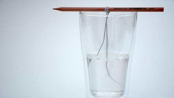 Glas mit Wasser, auf dem ein Bleistift mit Baumwollfaden liegt | Rechte: KiKA