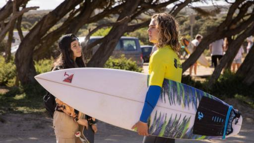 Das Mädchen Summer steht mit ihrem Skateboard gegenüber von Ari mit Surfbrett