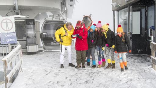 Schneewanderung in den Alpen. Singa, Volker Rosin und die TanzAlarm-Kids marschieren munter durch das verschneite Gelände und sehen nicht, wie sich der TanzTapir, angelockt von beschwingter Hüttenmusik, von der Truppe entfernt.