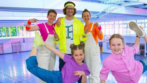 Die TanzAlarm-Kids stehen in sportlichen Posen vor Kindermusiker herrH und seinen Tänzerinnen mitten in der Sporthalle. TanzTapir steht rechts an der Seite mit einem Kletterseil in der Hand.