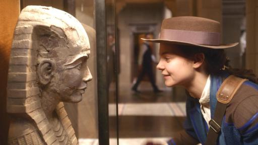 Im Museum: Theodosia (Eloise Little) schaut lächelnd und leicht zu ihr hin gebeugt, einer steinernen Pharaonen-Statue in die Augen. Sie trägt einen braunen Hut.