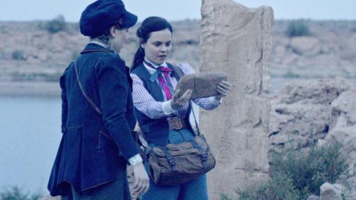 Maya und Theo finden in einer ägyptischen Ruine einen Hinweis auf einem Stein. Sie stehen nebeneinander und Theo hält den Stein mit beiden Händen.