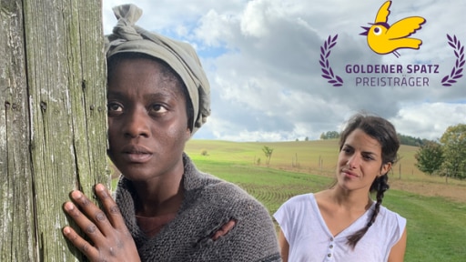 Clarissa steht hinter Harriet Tubman (Nancy Mensah-Offe) auf einem Feld. Tubman lehnt mit Körper und Gesicht an einen Holzphal. Sie schaut lauschend geradeaus. Clarissa beobachtet sie.