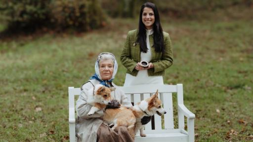 Königin Elisabeth II. sitzt mit ihren zwei Corgis auf einer Parkbank. Clarissa steht mit ihrer Zeitmaschine dahinter und lächelt.