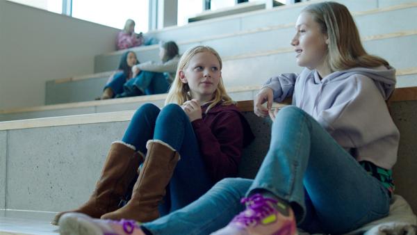 In einer Turnhalle - hell erleuchtet vom Tageslicht, sitzen Sanne (Tilde Ender Søvik) und Tara (Maya Ystenes) nebeneinander auf den hohen Stufen der Tribüne. Während Sanne erwartungsvoll zu Tara schaut, ist deren Blick zum Spielfeld gerichtet. Tara lächelt leicht. Beide tragen Jeans und Hoodie.