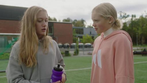 Bei Tageslicht stehen Tara und Mina sich gegenüber auf einem Wiesen-Sportplatz - im Hintergrund ist das Schulgebäude. Mina guckt zweifelnd in Taras Gesicht, während Tara ernst nach unten schaut, also ob sie Minas Blick ausweicht.