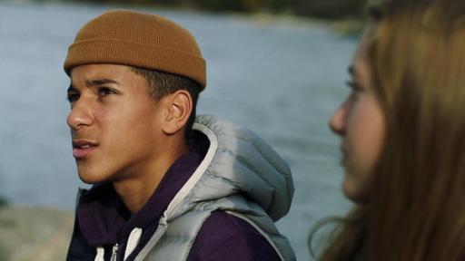 Bei Tageslicht sitzen Jonas und Tara nebeneinander am Wasser. Jonas trägt eine dicke Jacke zusätzlich mit Daunenweste darüber und eine Mütze auf dem Kopf. Er schaut mit ernstem Gesicht seitlich nach oben, während Tara ihn anschaut.