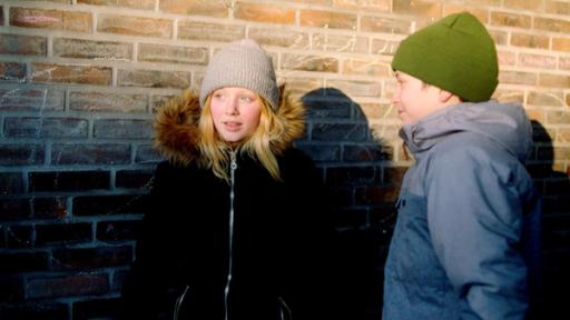 Daniel und Sanne stehen sich auf dem Schulhof gegenüber. Daniel lächelt Sanne an und sie schaut verlegen zur Seite. Sie sind in dicke Jacken und Mützen eingepackt.